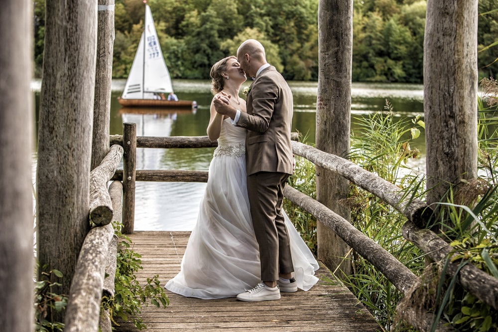 Ein Hochzeitspaar umarmt sich auf einem Holzsteg.Im Hintergrund ist Wasser zu sehen. Ein kleines Segelschiff fährt im, Hintergrund vorbei.