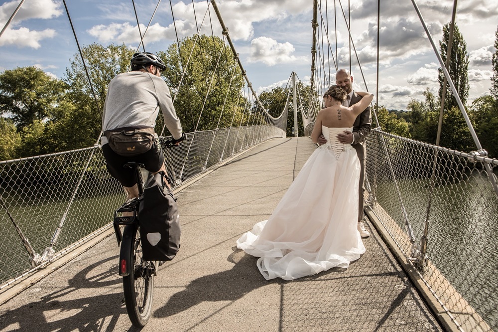 Ein Hochzeitspaar steht auf einer Brücke und umarmt sich. Ein Fahrradfahrer fährt an Ihnen vorbei