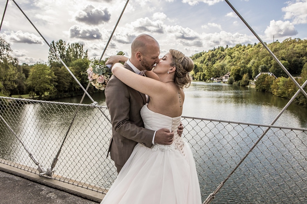 Hochzeitspaar umarmt und küsst sich auf einer Brücke. Im Hintergrund ist ein Fluss und grüne Büme zu sehen