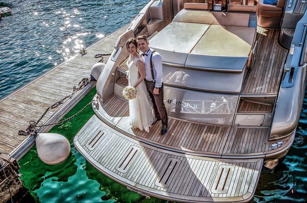 Hochzeitspaar steht auf einer Yacht.Von oben fotografiert. Beide schauen in die Kamera.