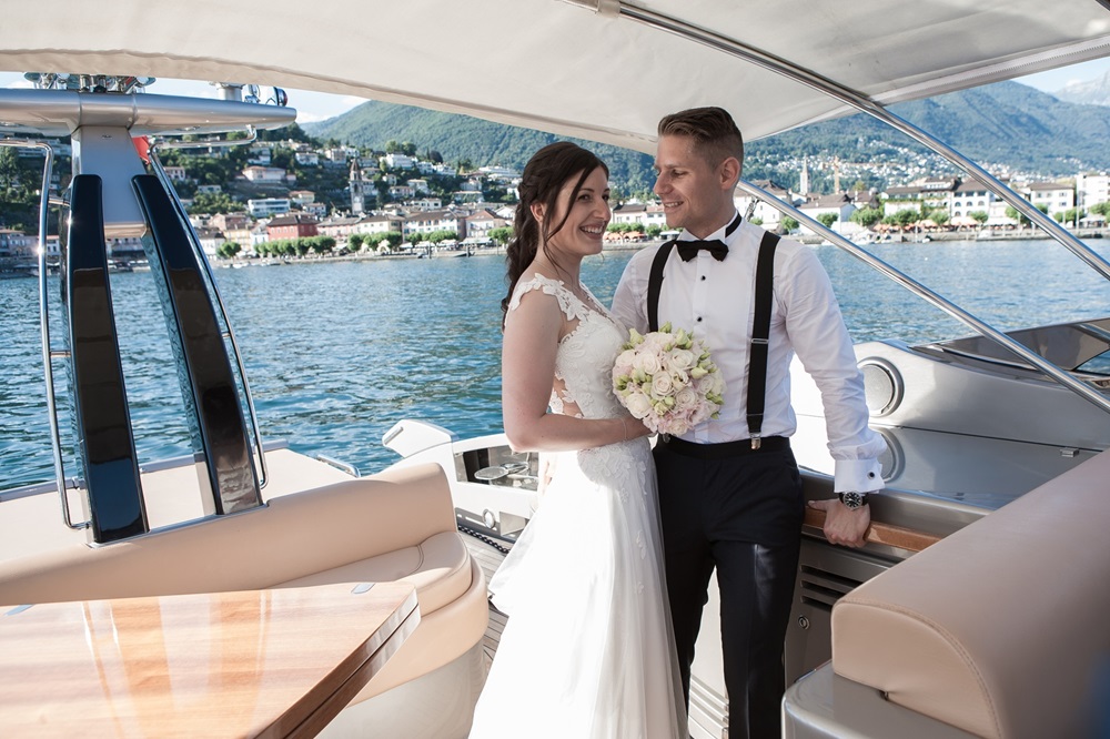Ein Hochzeitspaar steht auf dem Deck einer Yacht. Sie stehen unter einem Stoffdach, im Hintergrund ist eine Stadt zu sehen.